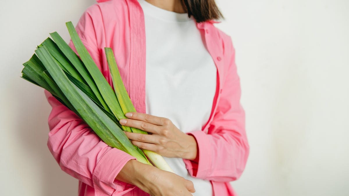 A woman in a pink jacket holding fresh spring onions against a white background.